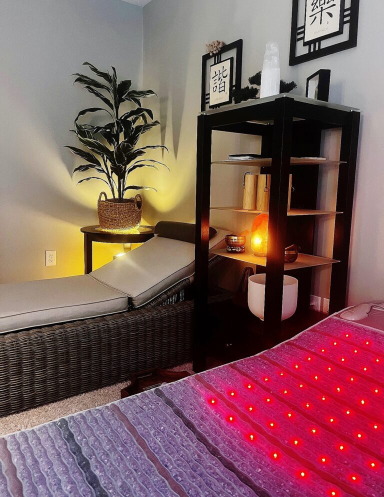 A tranquil looking spa like room containing a PEMF wellness mat lined with amethyst, tourmaline, obsidian and jade displays illuminated Red light therapy lights. Nearby, a linen cushioned rattan chaise lounge with bolster and a black glass shelved bookcase containing a Crystal Singing bowl and Himalayan singing bowls, along with Koshi chimes furnish this Zen looking sound therapy space.